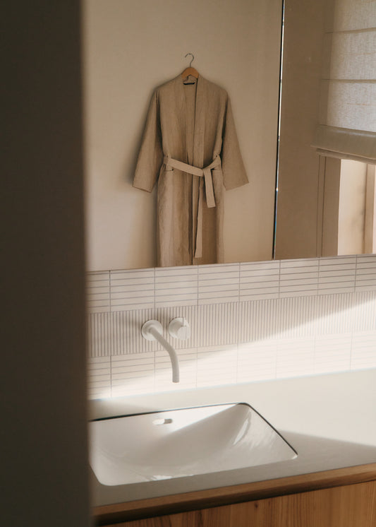 Unisex outerwear bathrobe, 100% Belgian linen, flax color, kimono-inspired with belt, hanging in a bathroom.