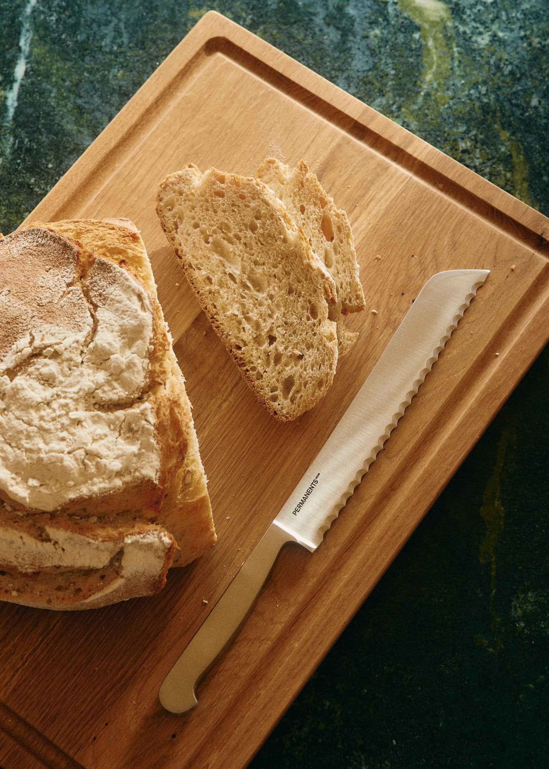 Silver steel PERMANENTS® bread knife with serrated blade on a wooden cutting board next to sliced artisan bread.