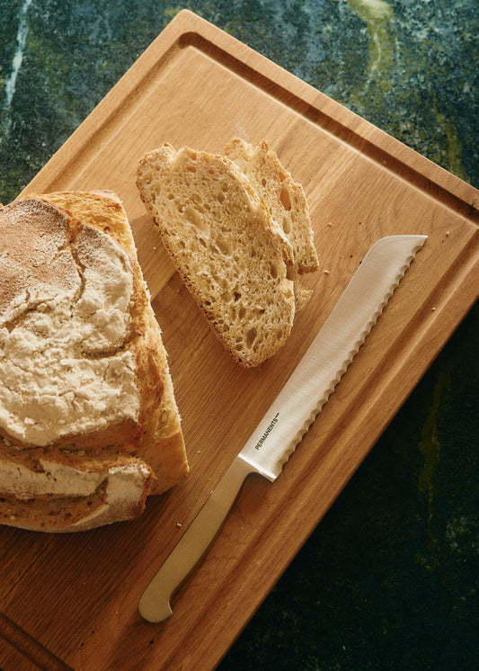 Silver steel PERMANENTS® bread knife with serrated blade on a wooden cutting board next to sliced artisan bread.
