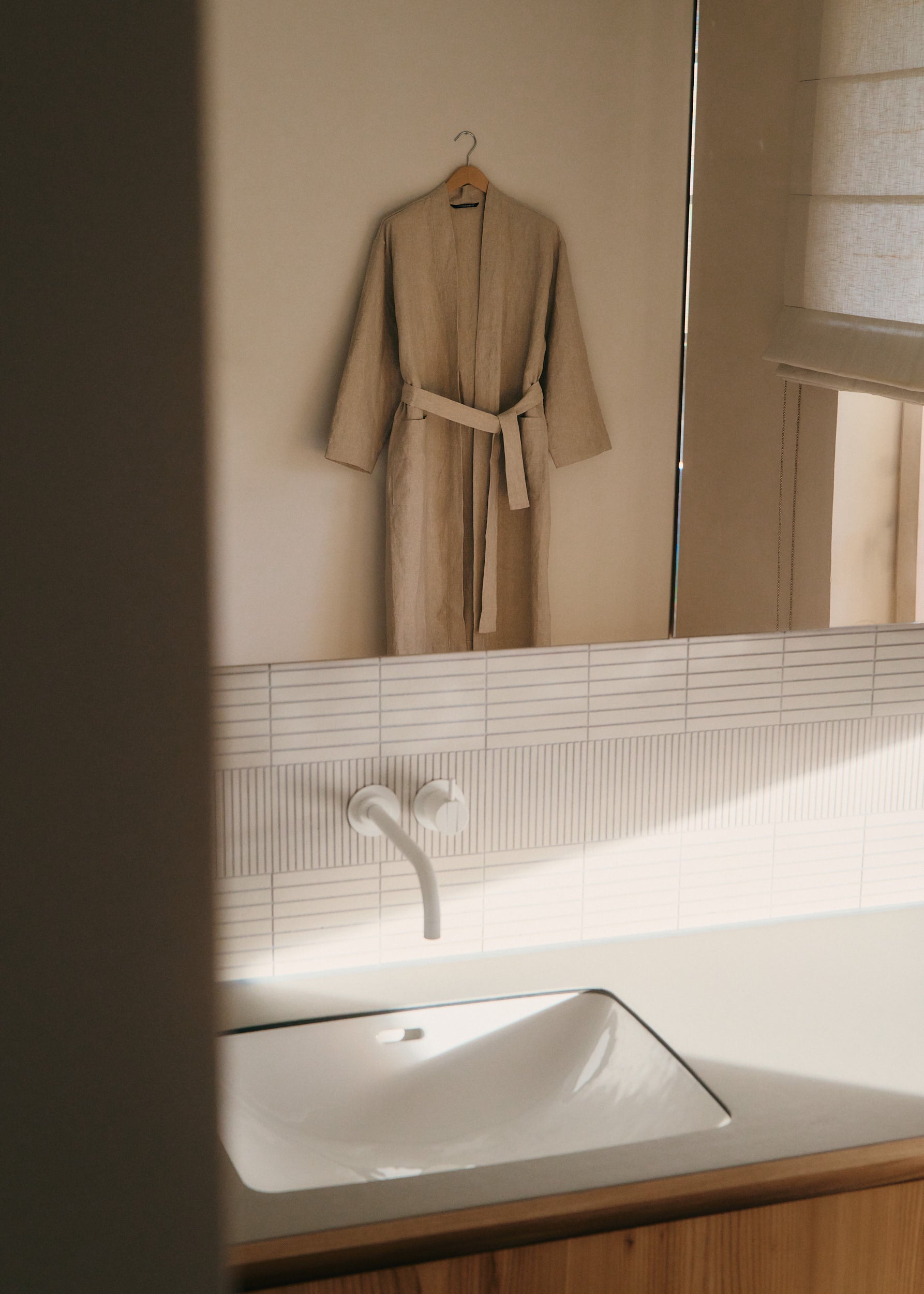 Unisex outerwear bathrobe, 100% Belgian linen, flax color, kimono-inspired with belt, hanging in a bathroom.