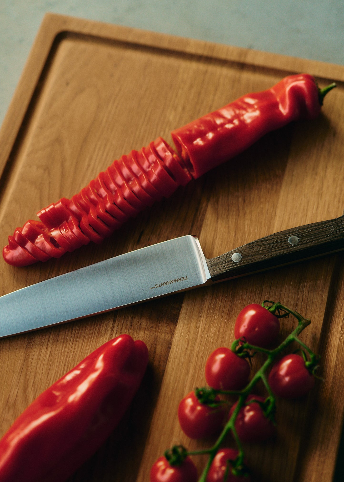 Chef's knife with a brushed steel blade and dark bog oak handle, next to sliced red pepper and cherry tomatoes on a wooden board.