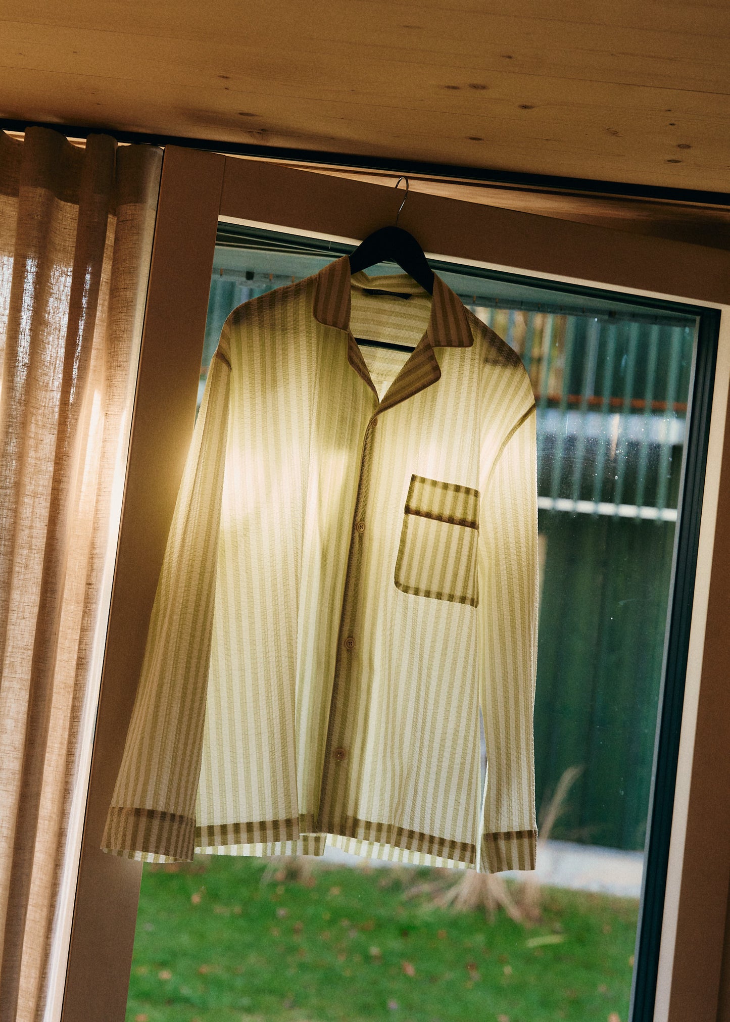 Unisex long-sleeved shirt, 100% organic cotton seersucker, sage stripe, with piped trim, hanging in a window.