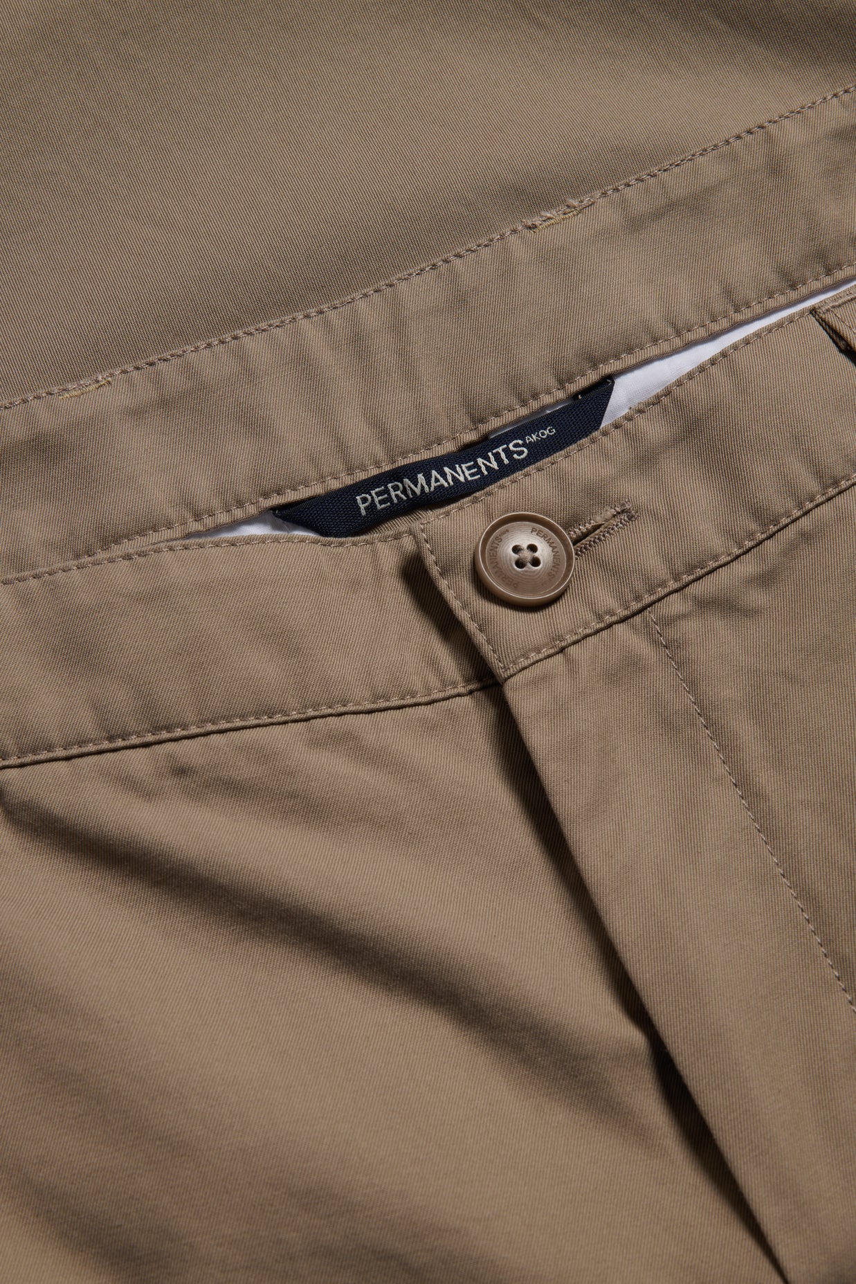Men's long-pants, organic cotton twill, washed beige, with button closure and "PERMANENTS" label.