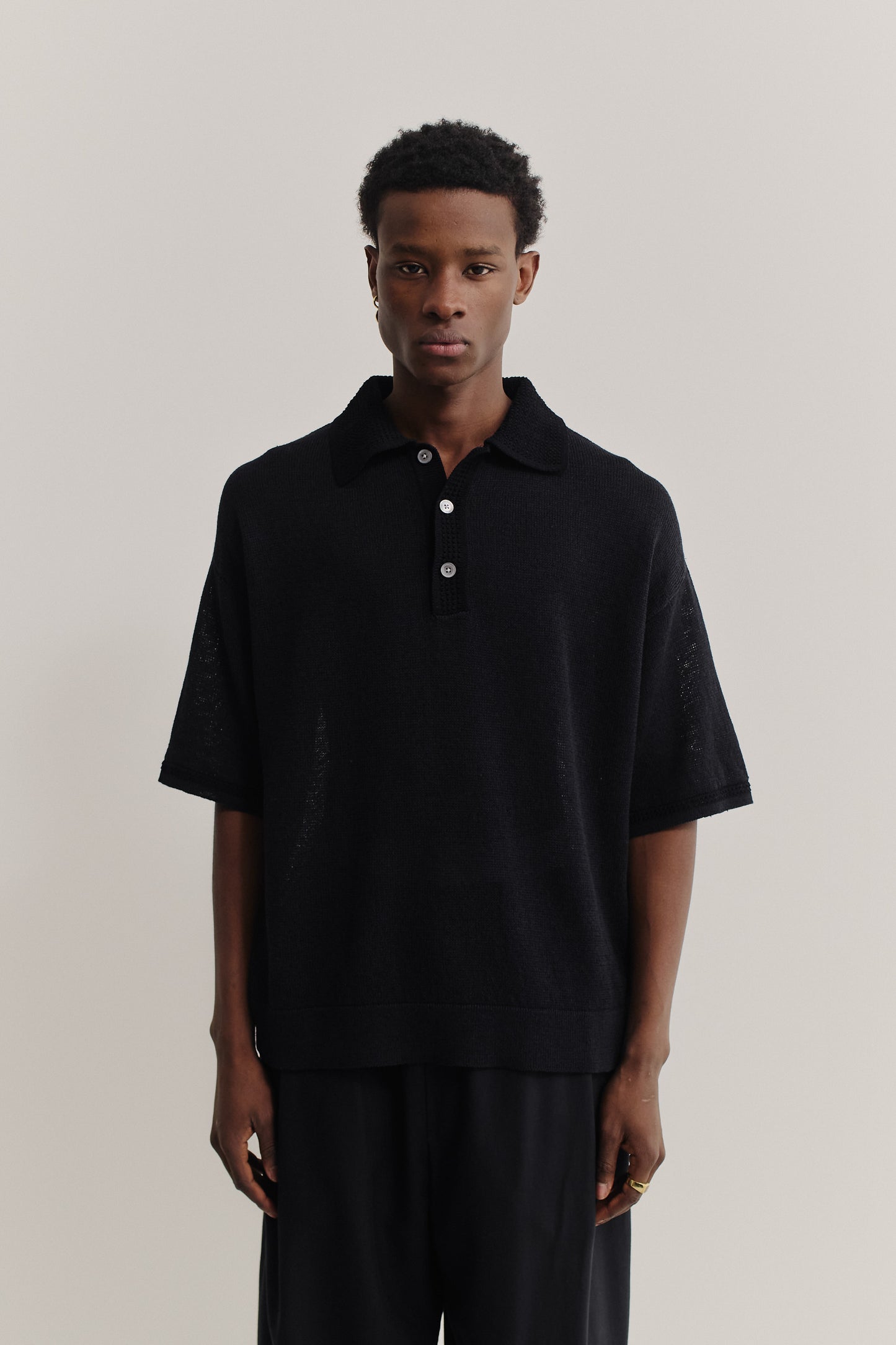 Men's knit short-sleeved polo, black linen-organic cotton blend, oversized fit, spread collar, white buttons.