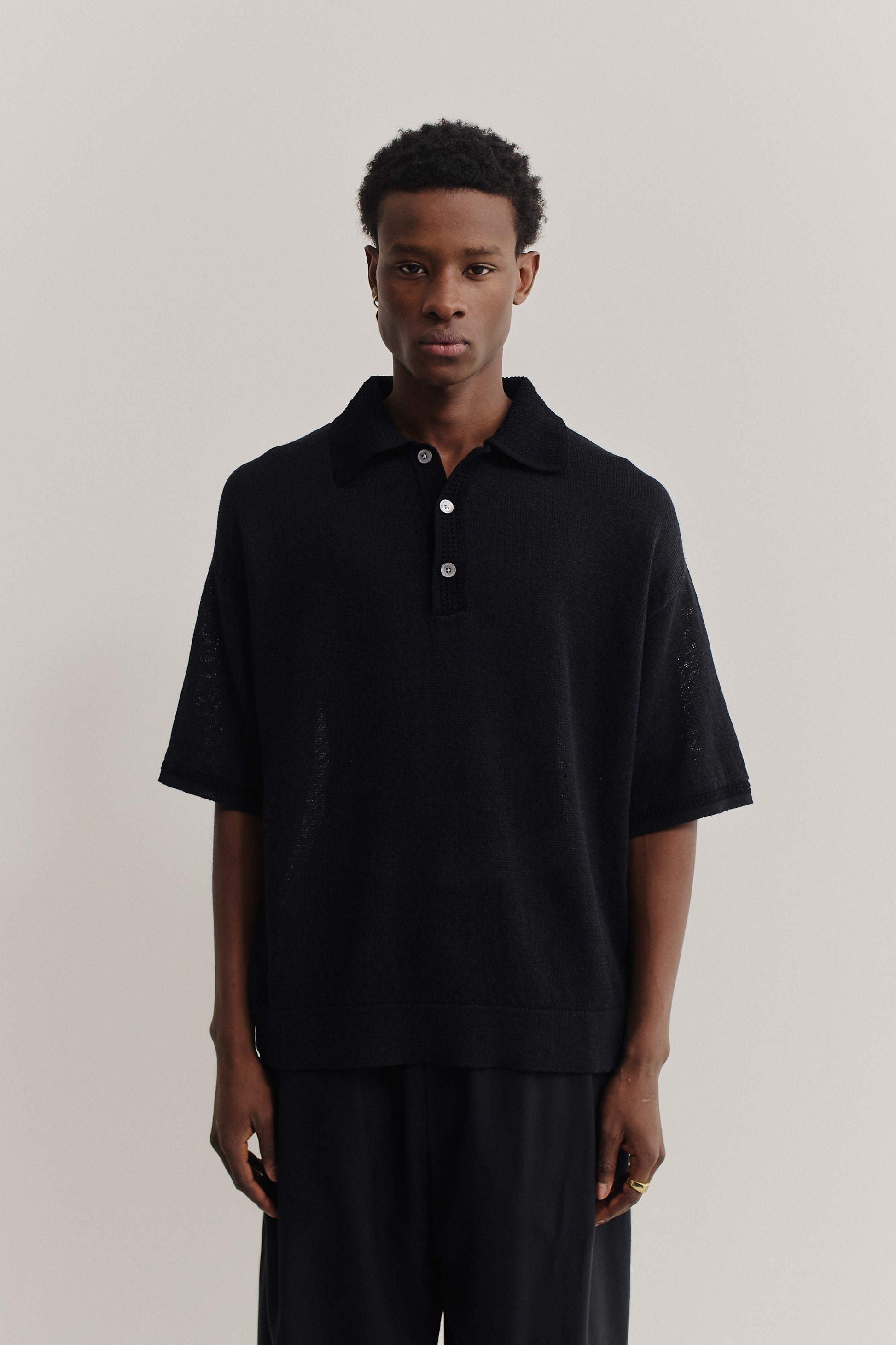 Men's knit short-sleeved polo, black linen-organic cotton blend, oversized fit, spread collar, white buttons.