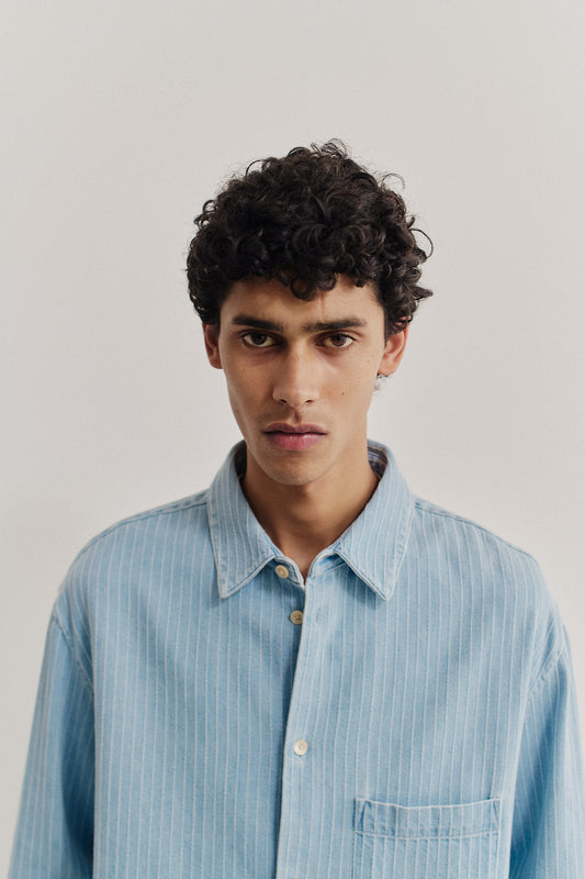 Men's long-sleeved shirt, organic cotton denim, washed light blue with subtle white pinstripes, oversized fit.