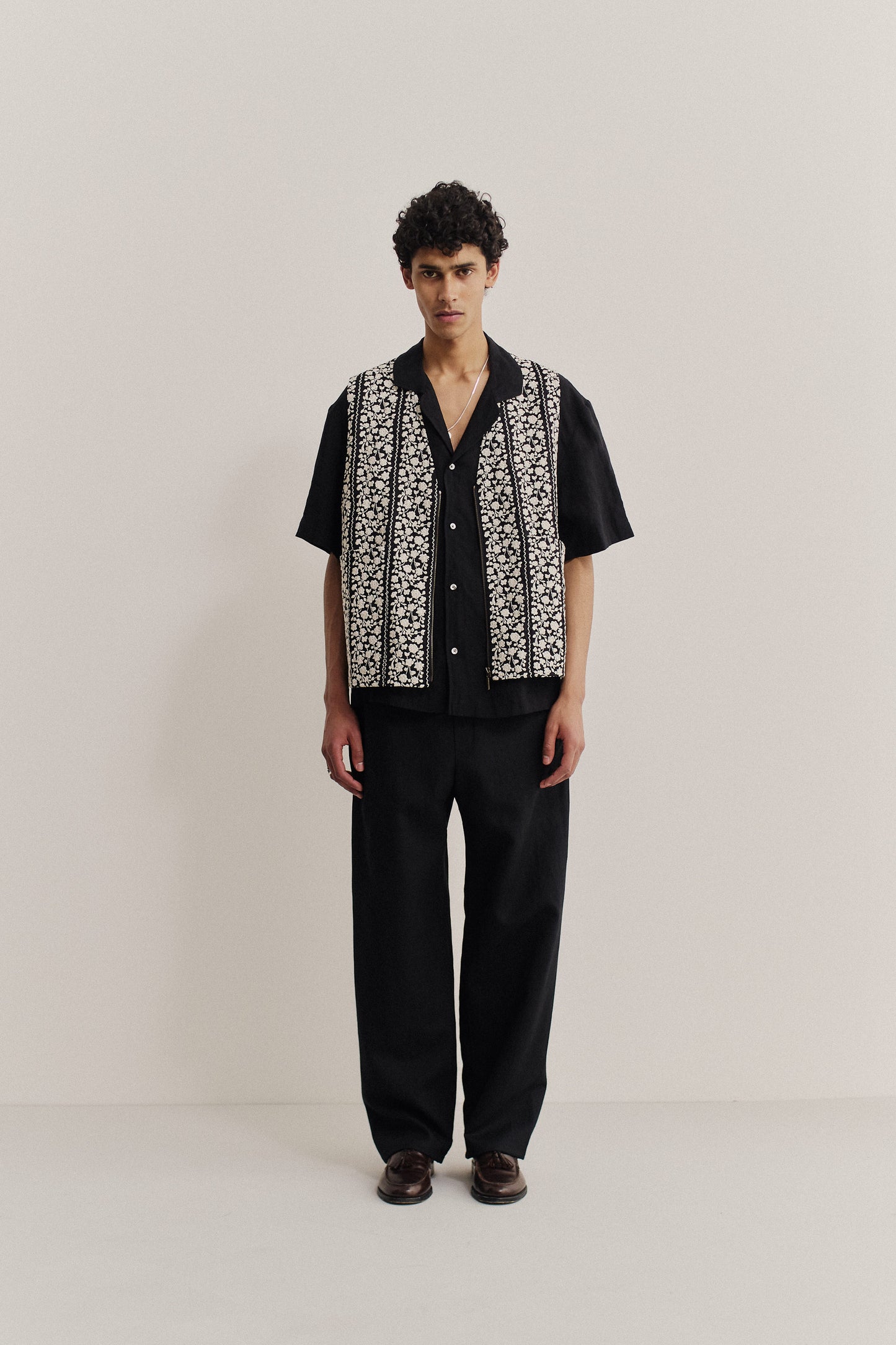 Men's outerwear vest, black cotton with white embroidered flower blocks, metal zipper, worn over a black shirt.