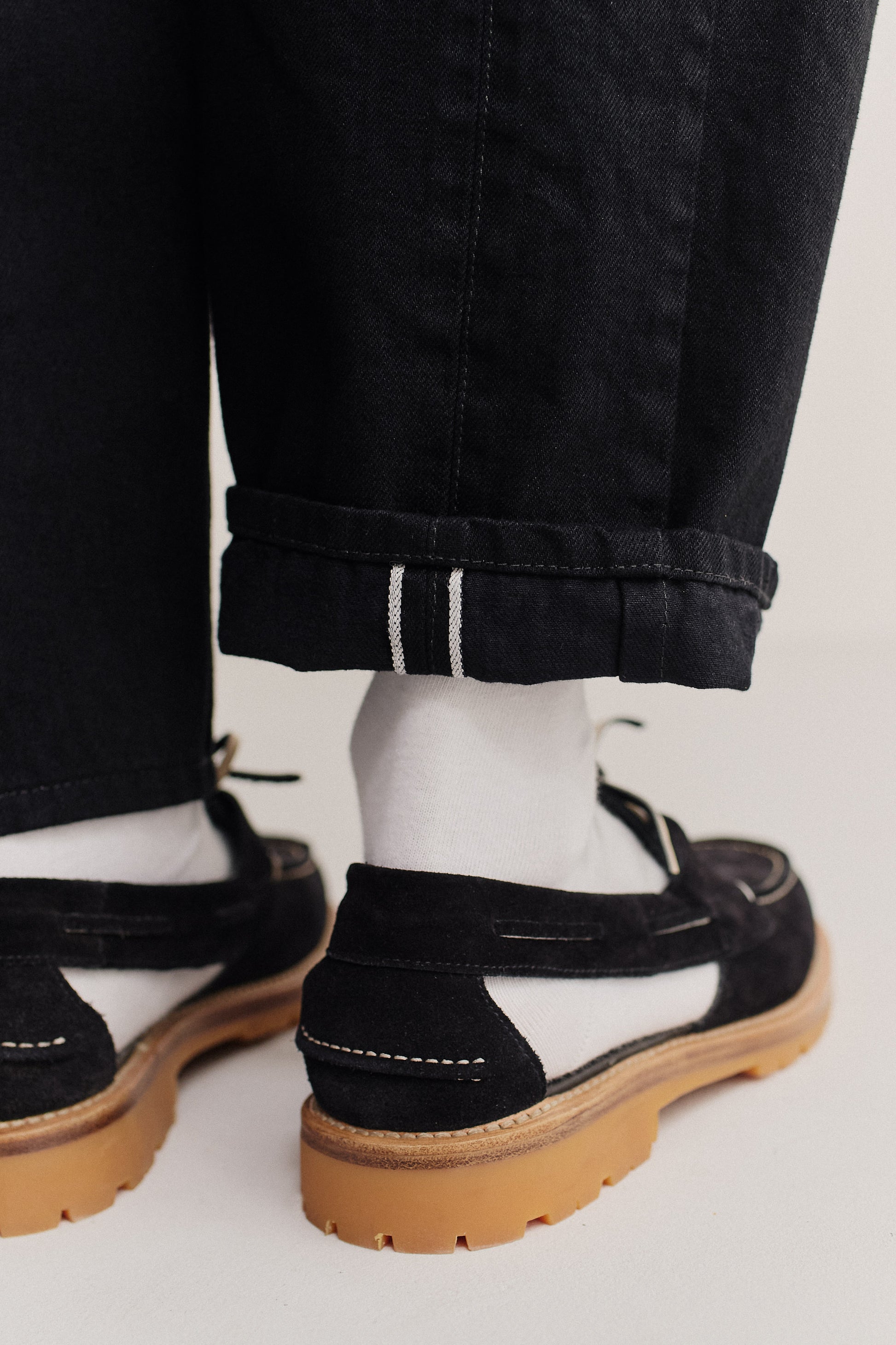 Men's long-pants, selvedge denim, black, with a cuffed hem revealing white selvedge detail, paired with black and white shoes.