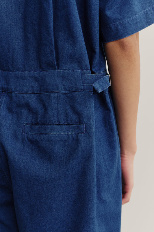 Women's jumpsuit, washed indigo cotton, short sleeves, adjustable buckle waistband, and patched back pocket.