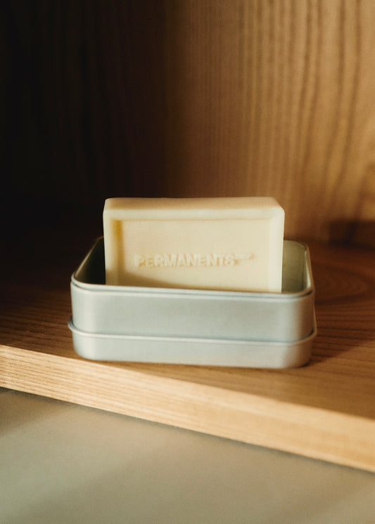 PERMANENTS® organic soap bar, off-white, in a silver aluminum travel tin on a wooden shelf.