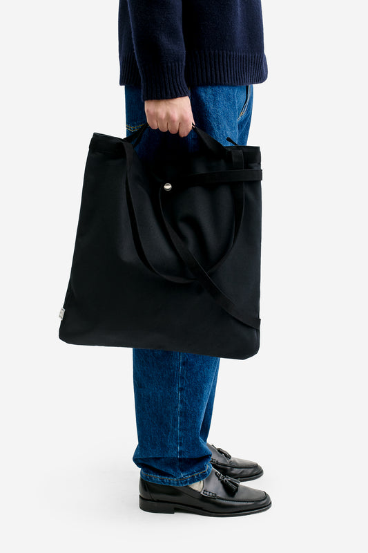 Men's accessories, black recycled cotton 3-Way Tote bag, held by hand, with adaptable strap and snap button.